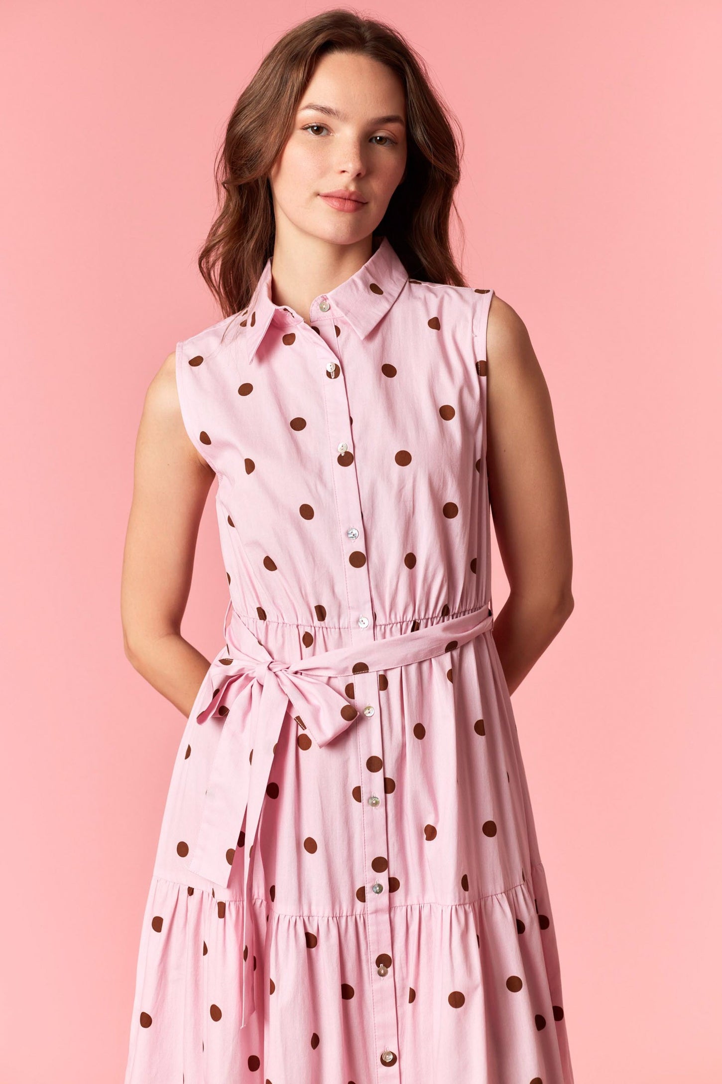 woman wearing a pink sleeveless button-up dress with brown polka dots and a tie belt detail
