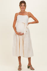 woman wearing a cream maternity dress with spaghetti straps, a tie at the waist, and holding her baby bump