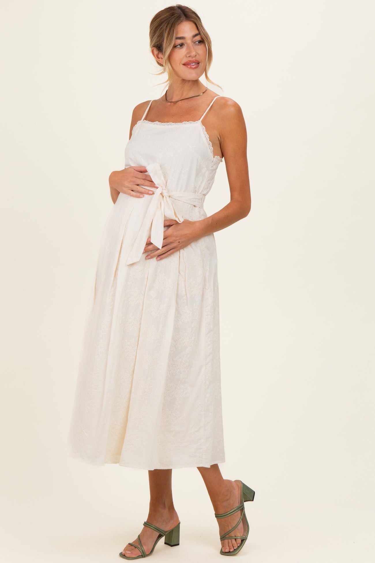 woman wearing a cream maternity dress with a tie waist and lace details, showing her baby bump