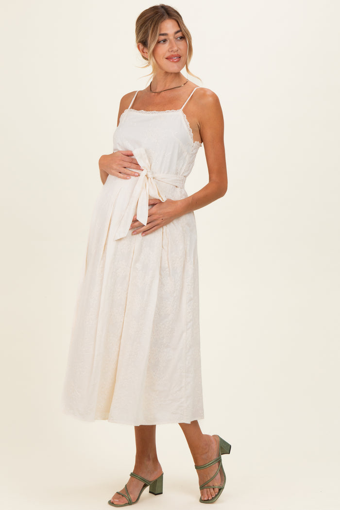 woman wearing a cream maternity dress with a tie waist and lace details, showing her baby bump