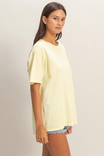 woman wearing a light yellow oversized t-shirt and distressed denim shorts, standing sideways with a relaxed pose