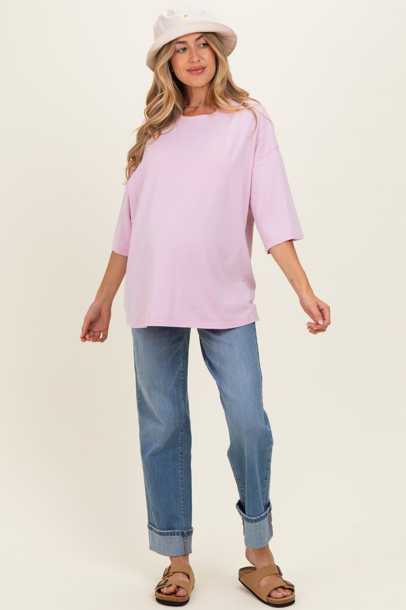 woman wearing a light pink oversized maternity top and blue rolled-up jeans, with a white bucket hat