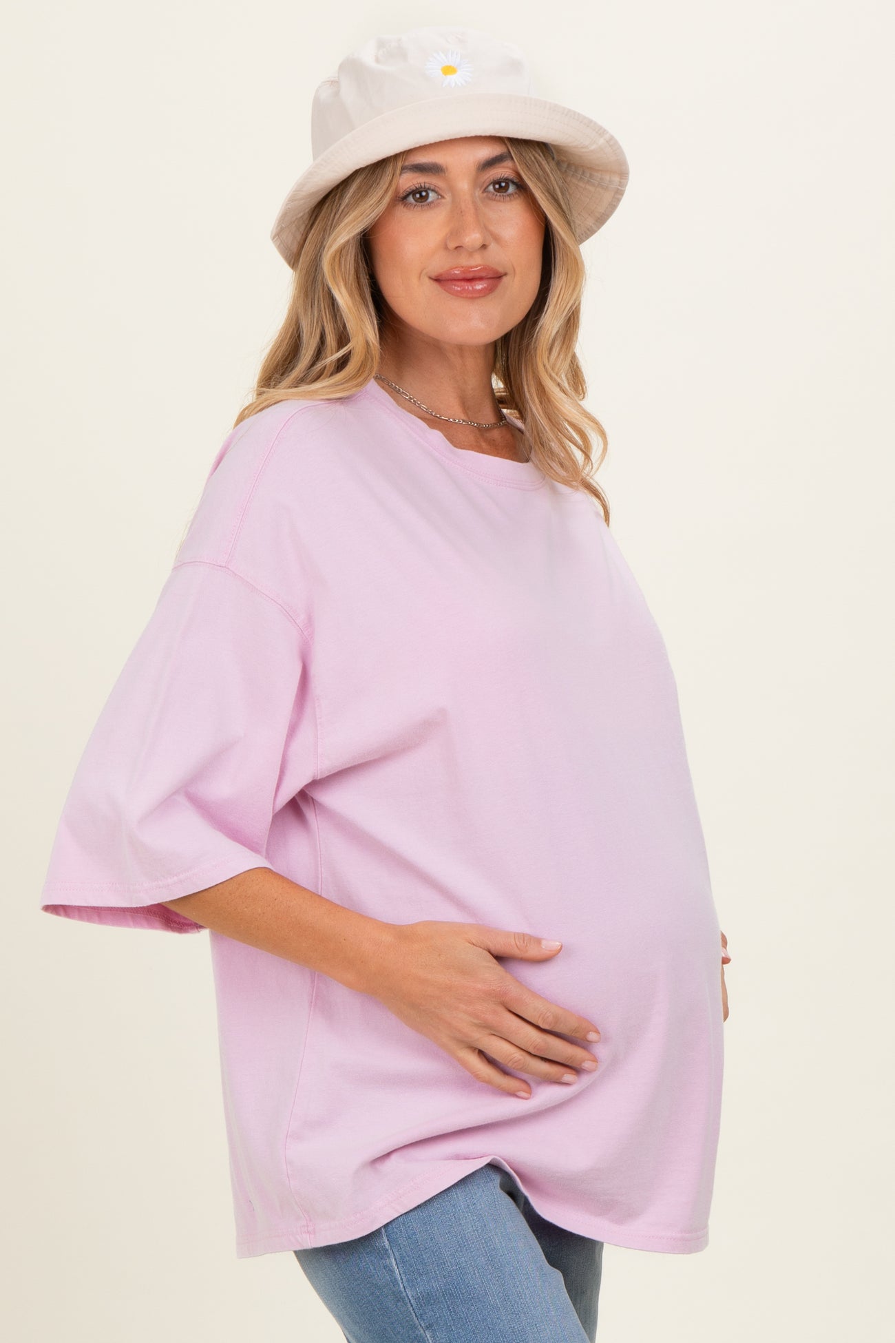 woman wearing a light pink oversized maternity t-shirt and holding her baby bump, styled with a cream bucket hat