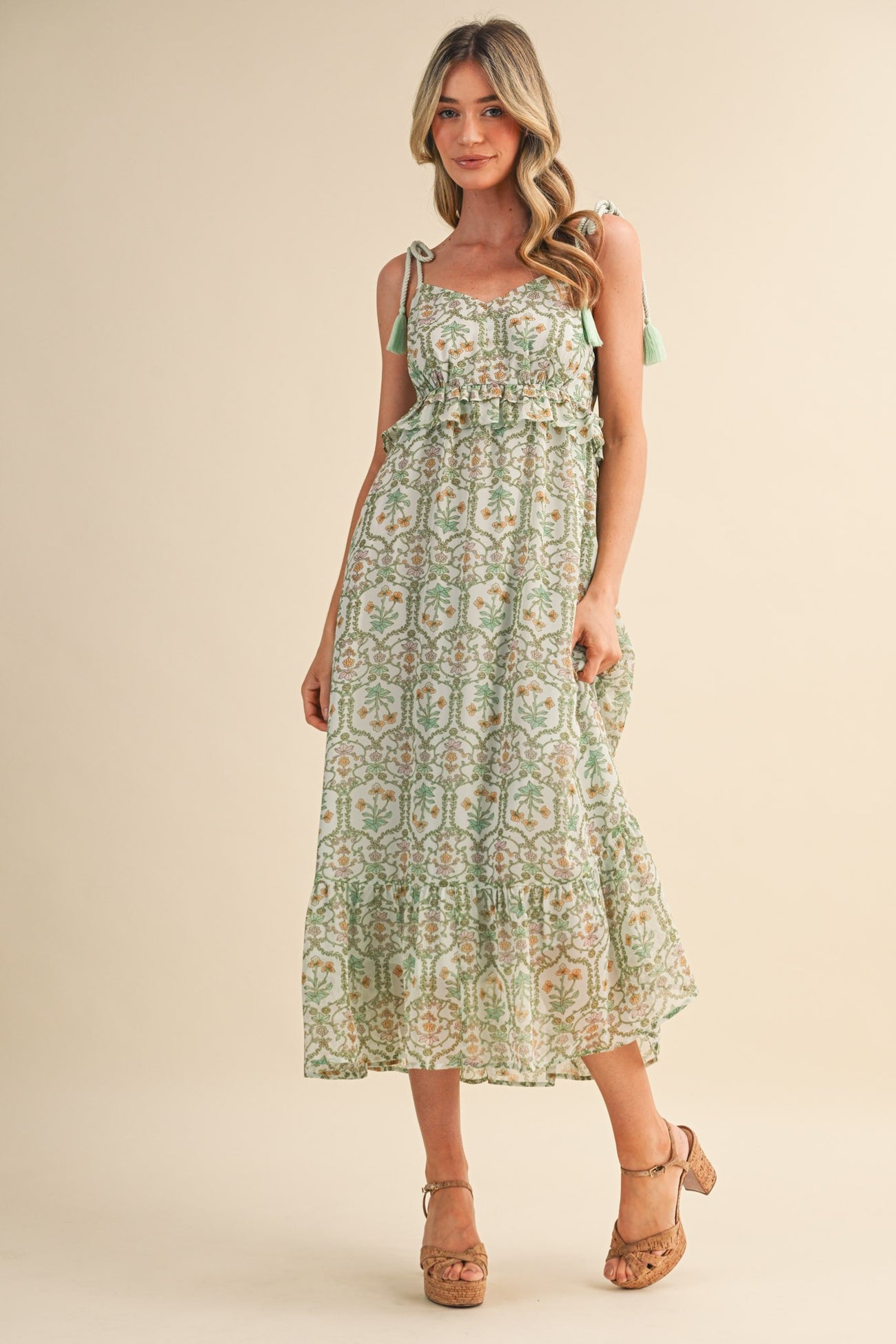 woman wearing a green floral maxi dress with ruffled details and tie straps, standing on a neutral background