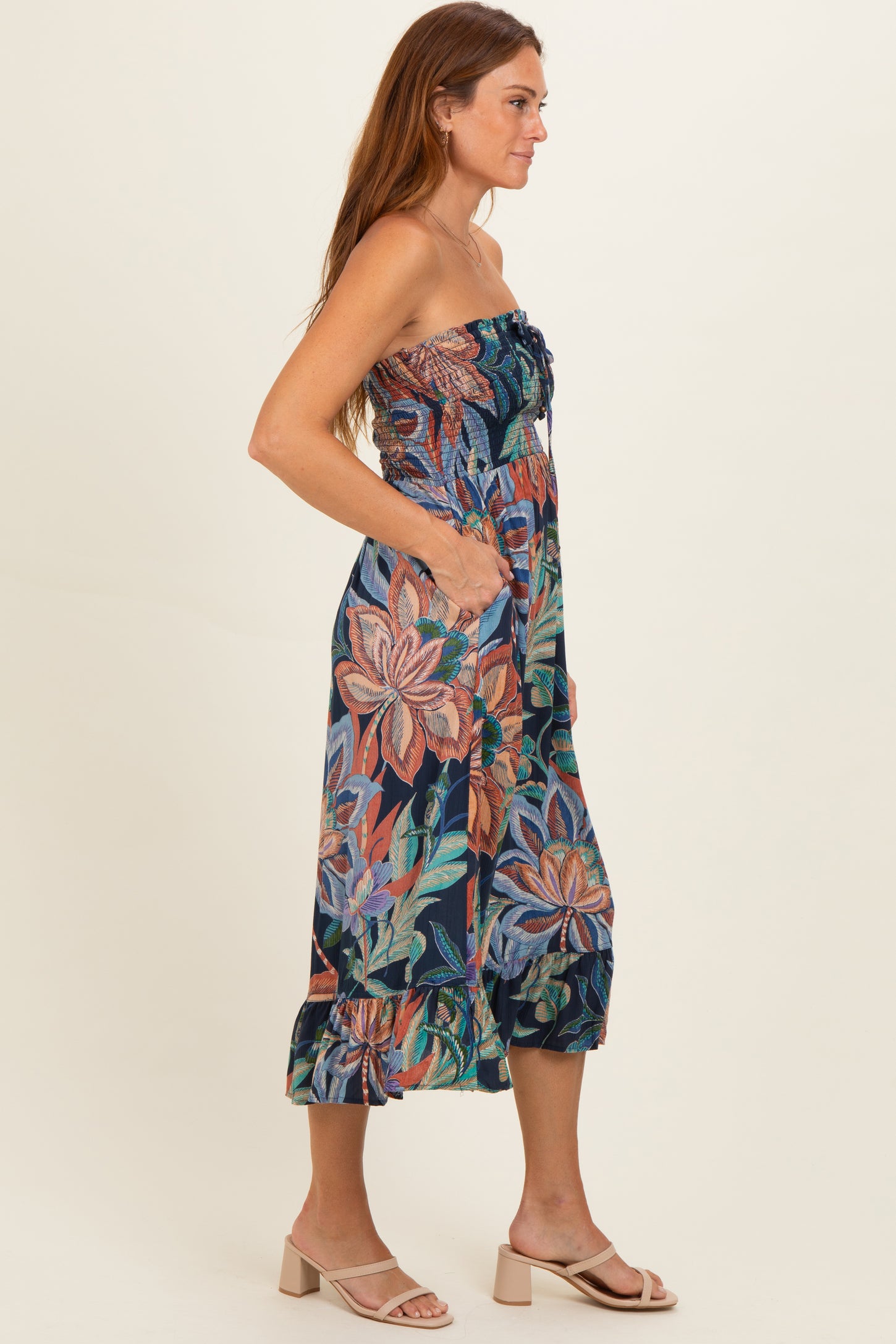 woman wearing a strapless floral maxi dress with pockets and ruffle hem, standing sideways against a beige background