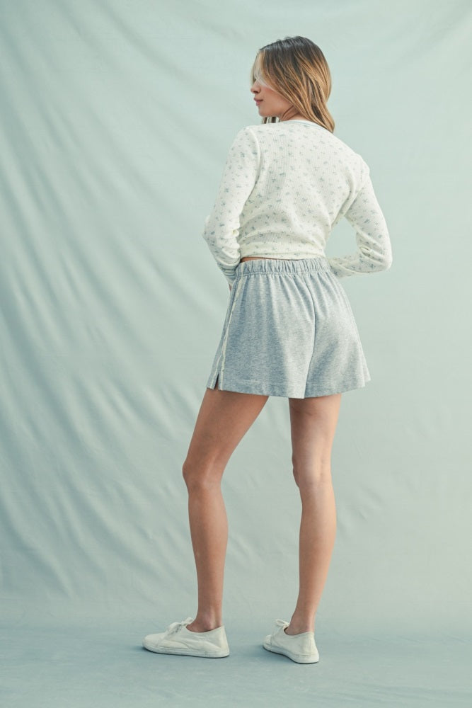 woman wearing a white cropped sweater with floral patterns and gray flared shorts, standing with her back to the camera