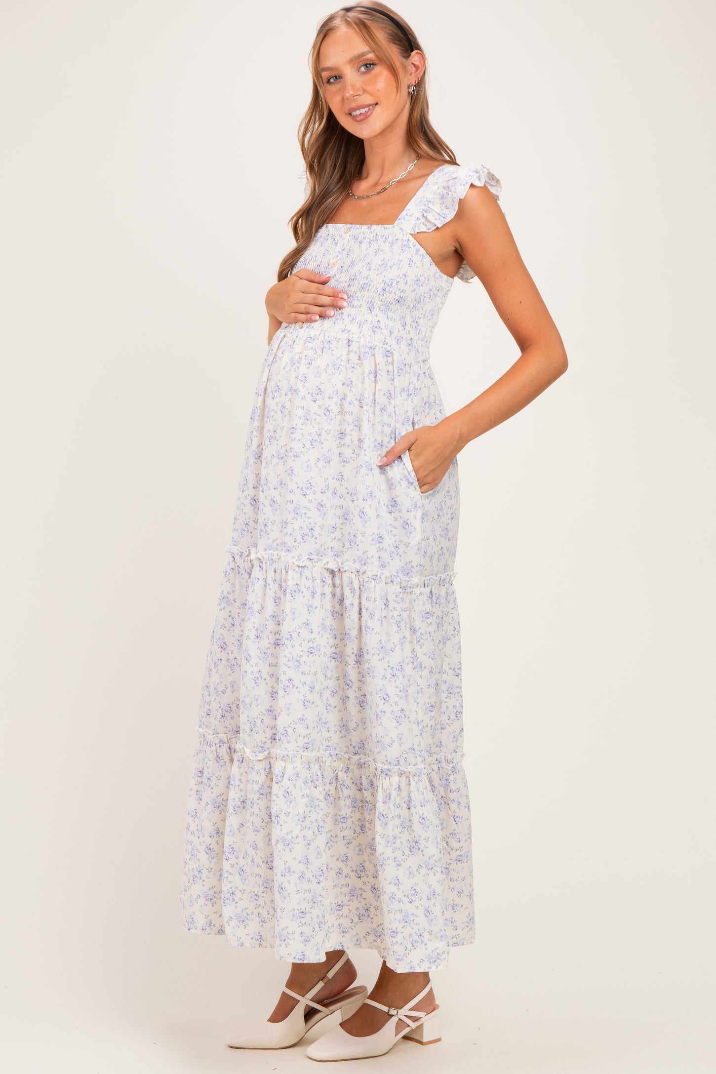 woman wearing a light blue floral maternity maxi dress with ruffle straps and pockets, holding her baby bump