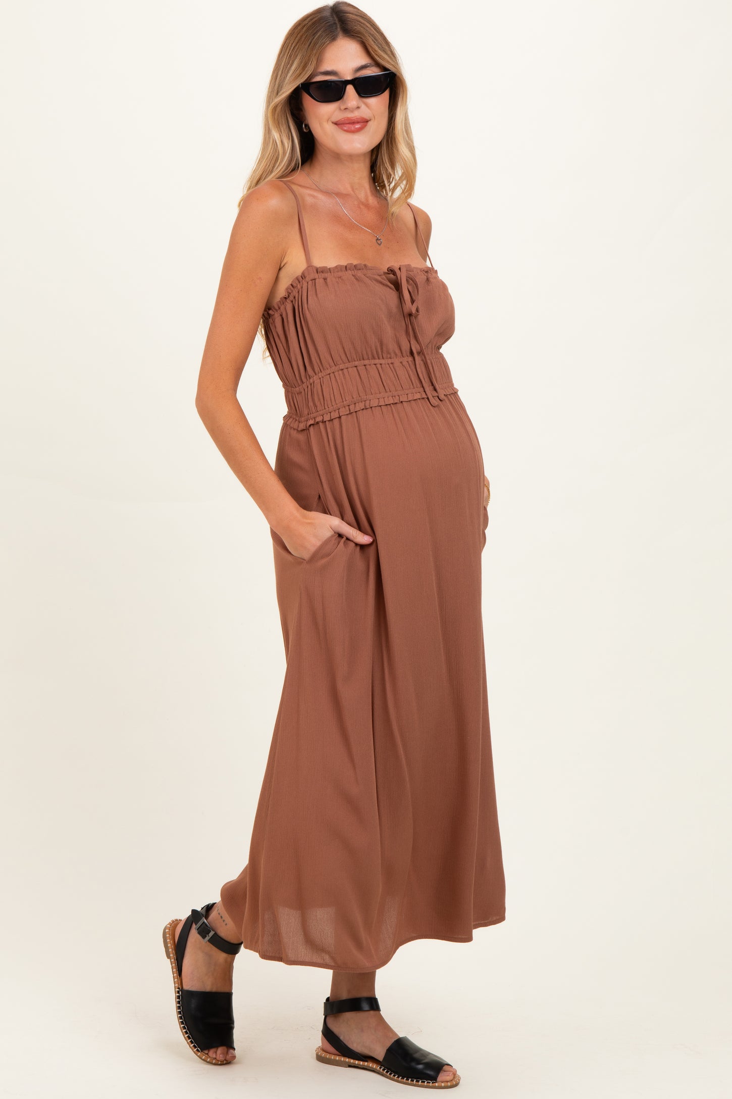 woman wearing a brown maternity maxi dress with adjustable straps and pockets, showcasing her baby bump