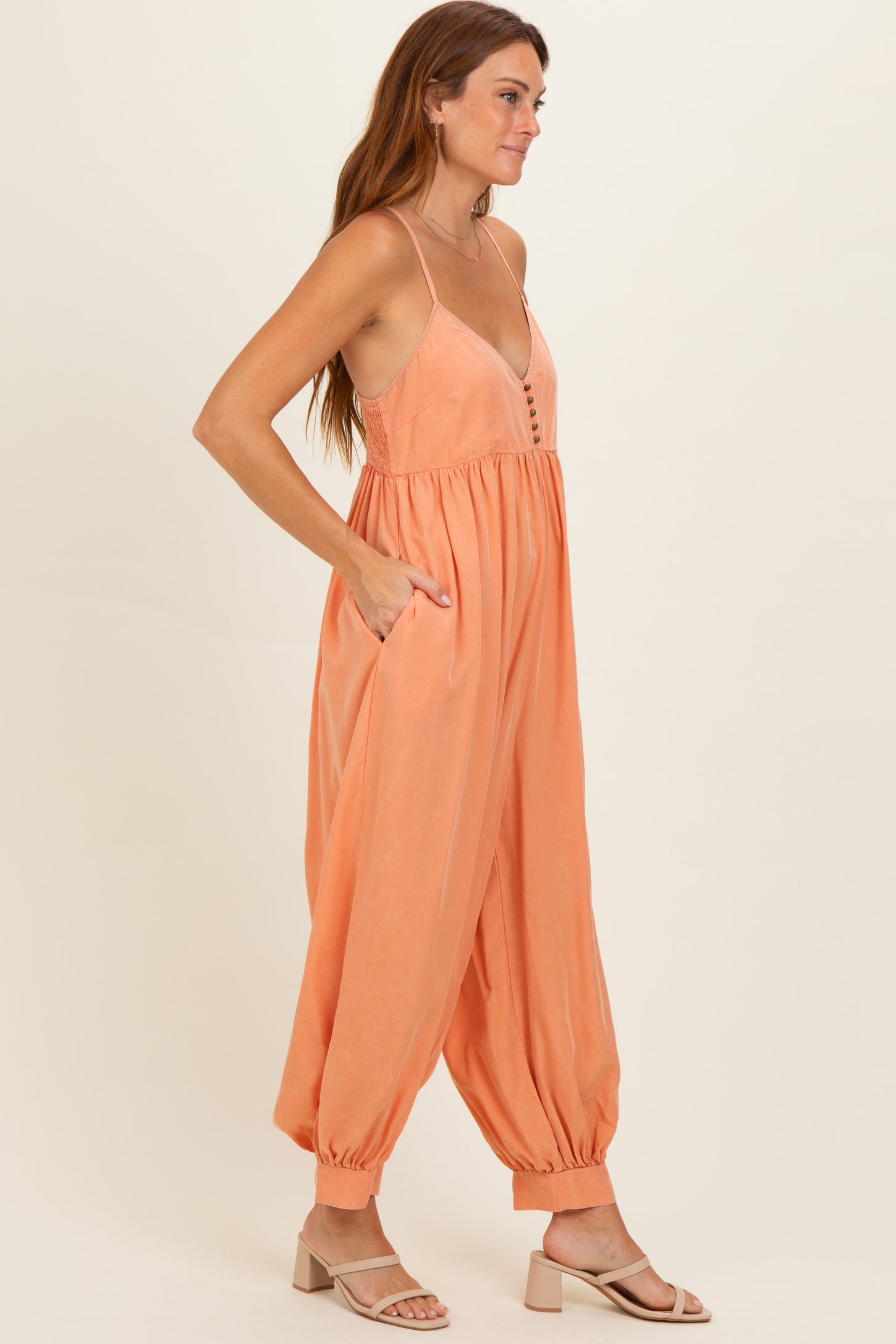 woman wearing a peach sleeveless jumpsuit with buttons and pockets, styled with beige sandals