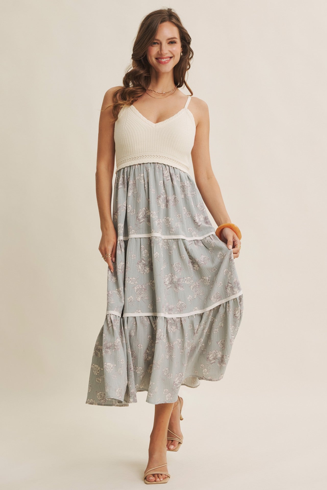 woman wearing a light grey floral maxi dress with a white bodice and tiered skirt, smiling and holding an orange