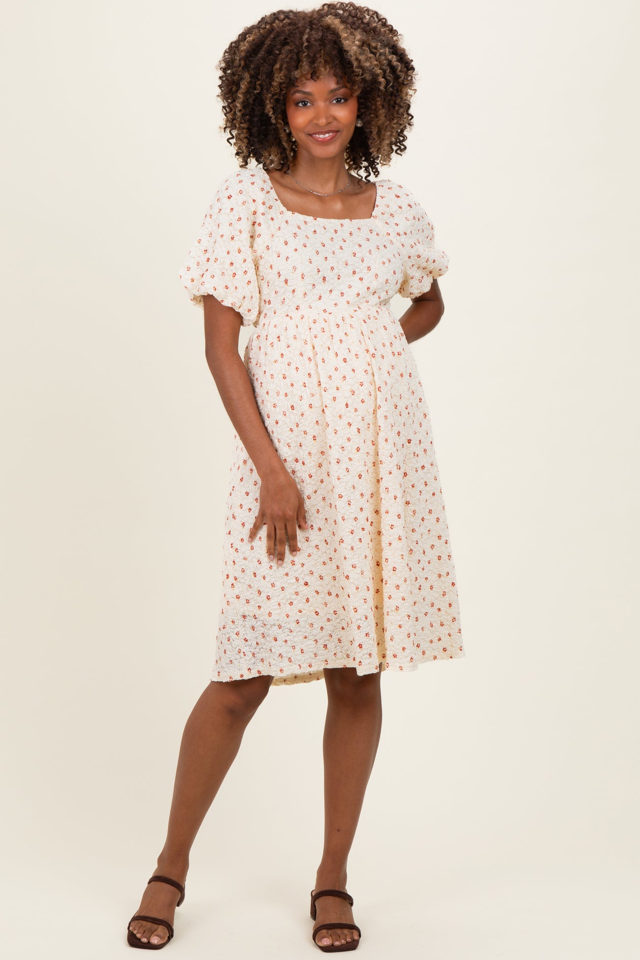 woman wearing a light yellow maternity dress with a square neckline and floral print, standing with a relaxed pose