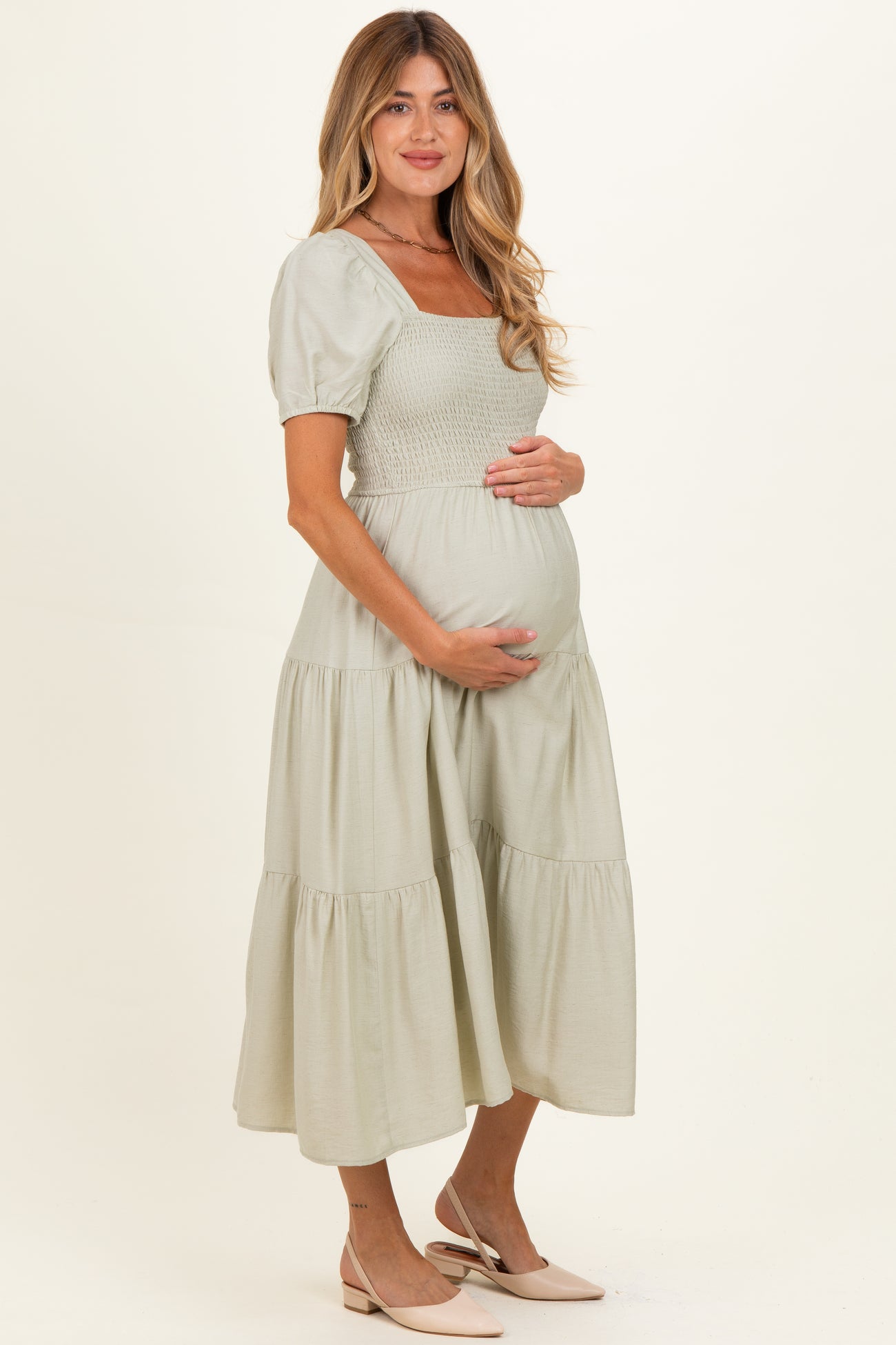 woman wearing a sage green maternity dress with short puff sleeves and a tiered skirt, holding her baby bump