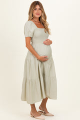 woman wearing a sage green maternity dress with short puff sleeves and a tiered skirt, holding her baby bump