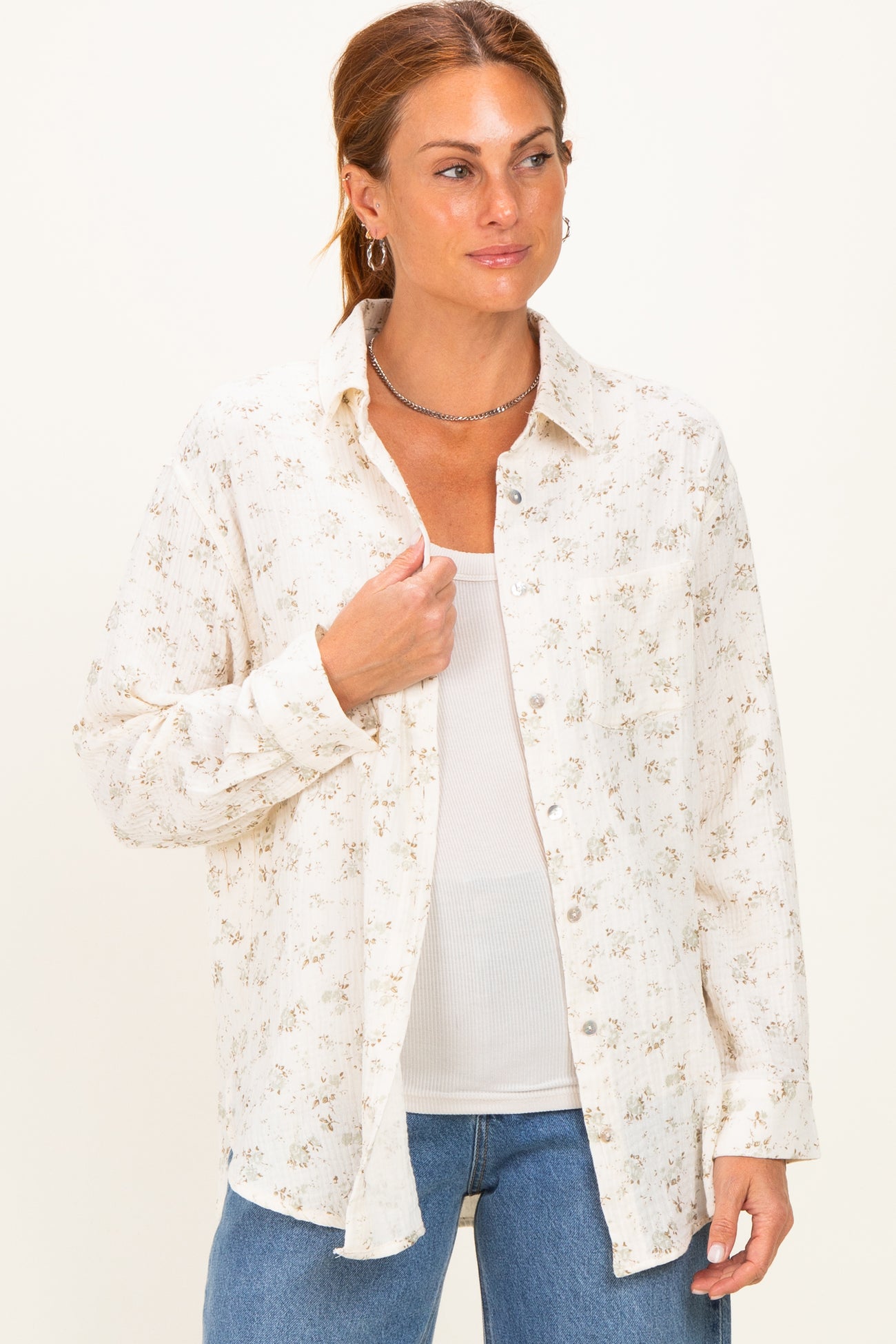 woman wearing a cream floral button-up shirt over a white tank top and blue jeans
