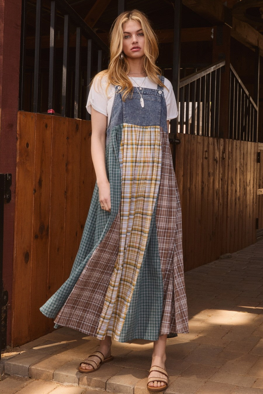 woman wearing a multi-patterned maxi dress with denim bib and plaid details, styled with a white t-shirt and sandals