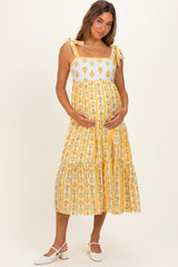 woman wearing a yellow floral maternity dress with tiered skirt and tie straps, holding her baby bump