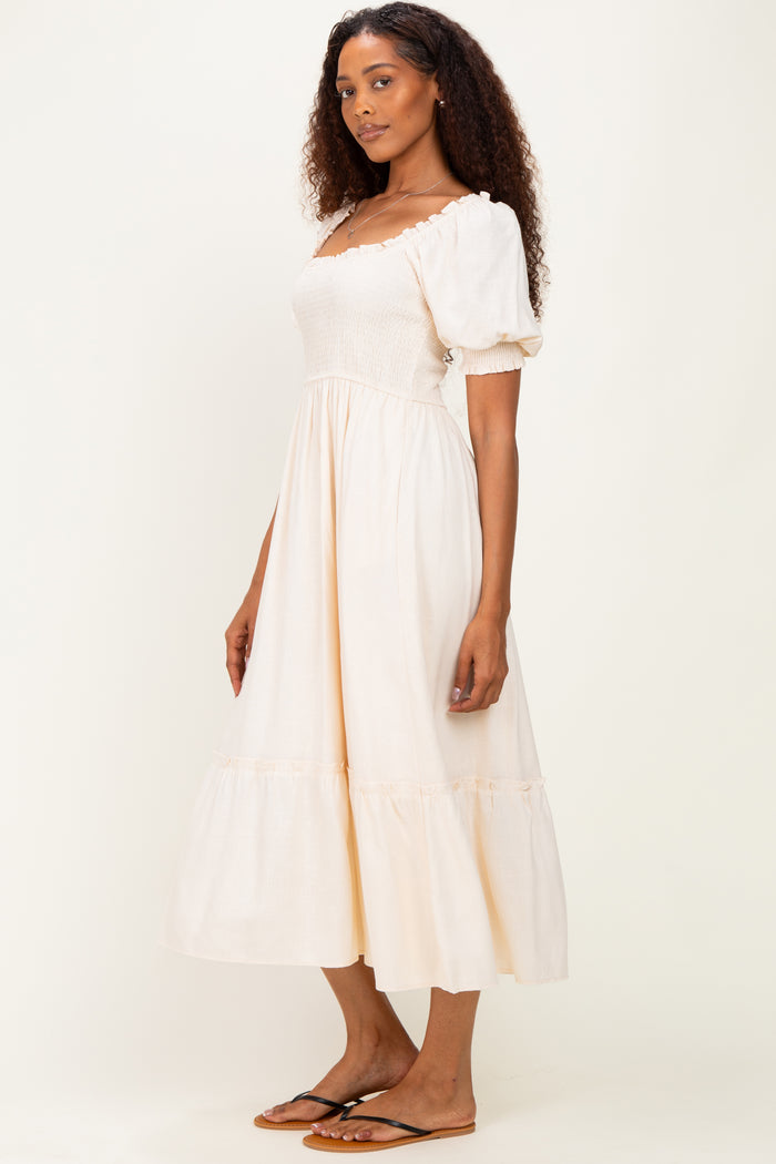 woman wearing a light beige smocked midi dress with puff sleeves and a tiered skirt, standing sideways