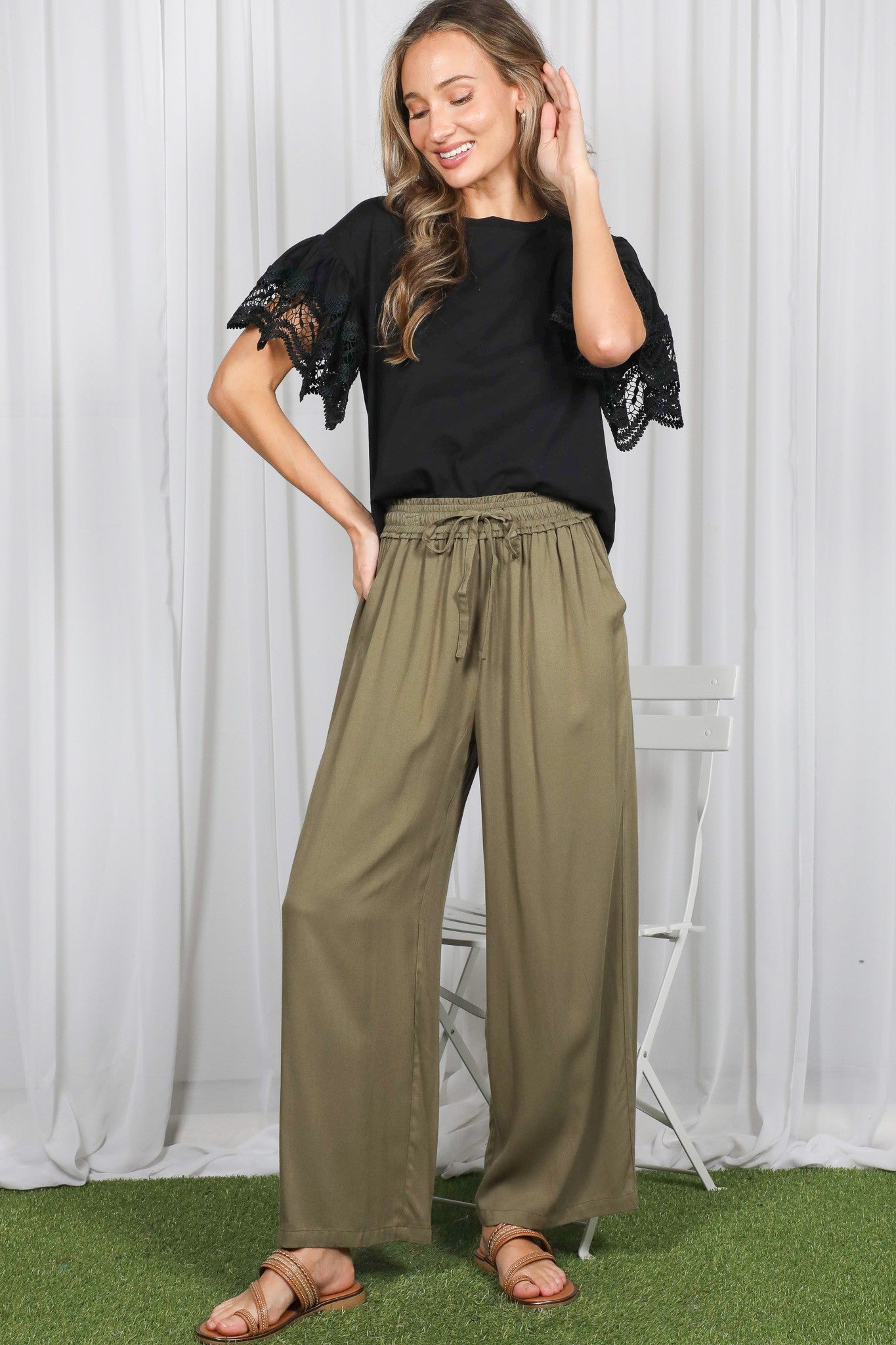 woman wearing a black top with lace sleeves and olive wide leg pants, standing on grass