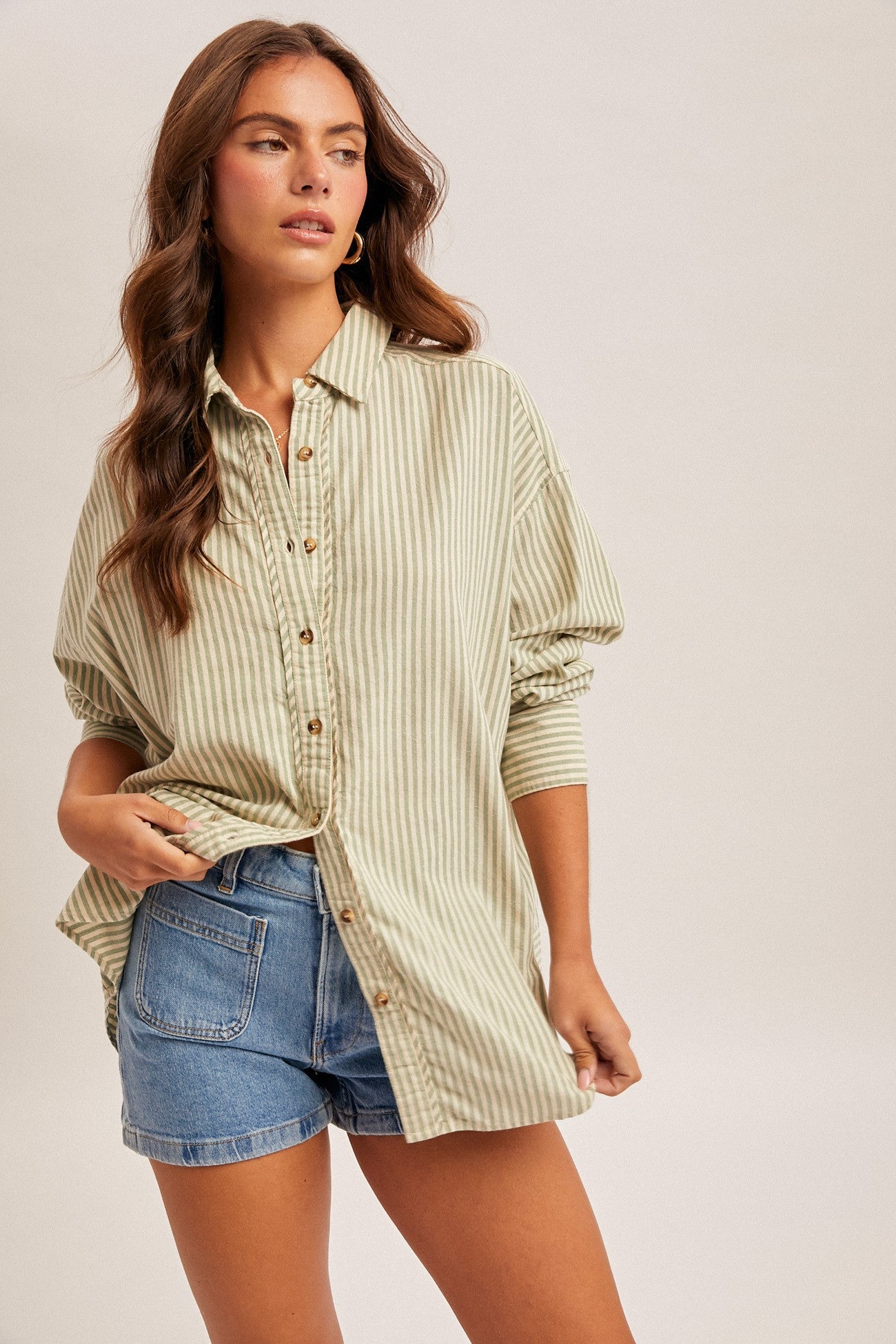 woman wearing a light green striped oversized shirt and denim shorts, posing casually