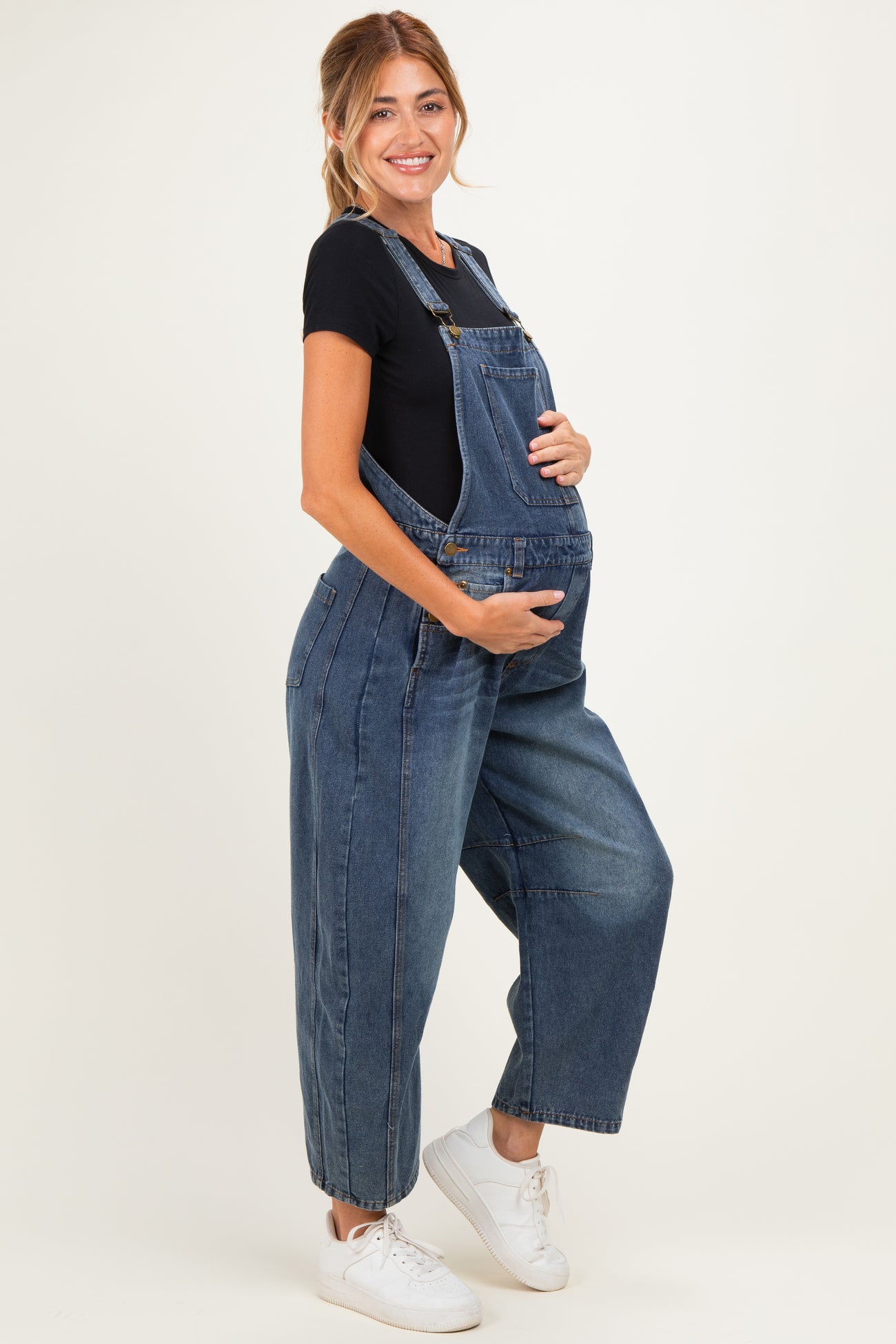 woman wearing a dark blue denim maternity overall, holding her baby bump with a black t-shirt underneath