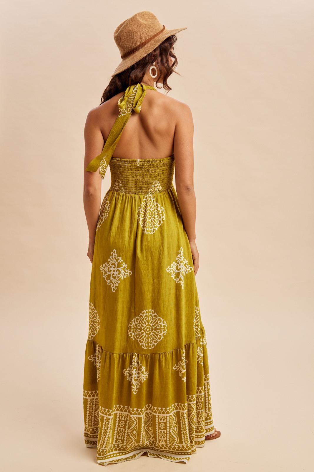 woman wearing a mustard yellow maxi dress with intricate white patterns and a halter neck tie, seen from the back