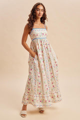 woman wearing a sleeveless floral maxi dress with blue straps and pockets, standing on a neutral background