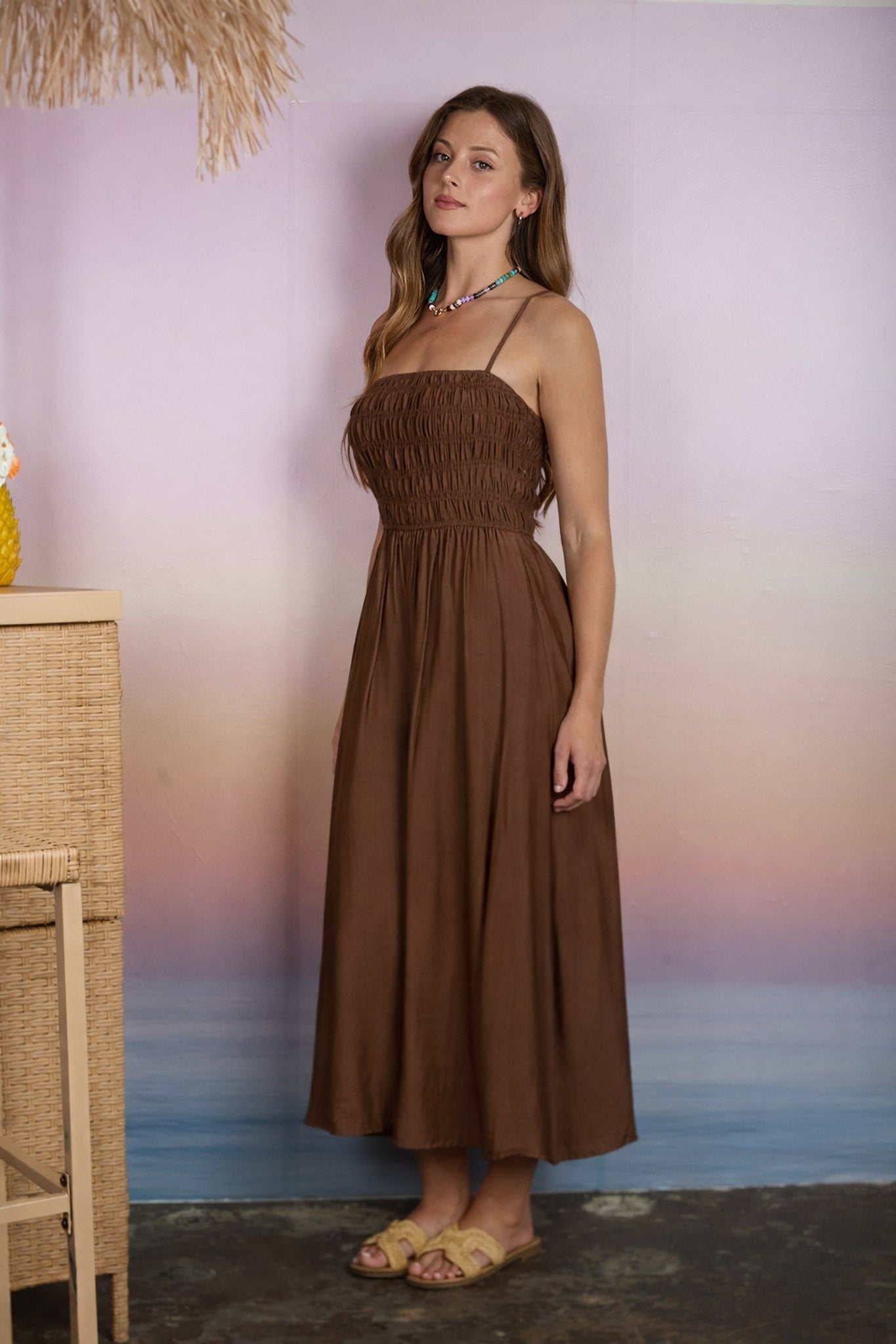 woman wearing a brown strapless maxi dress with smocked bodice, standing against a pastel background