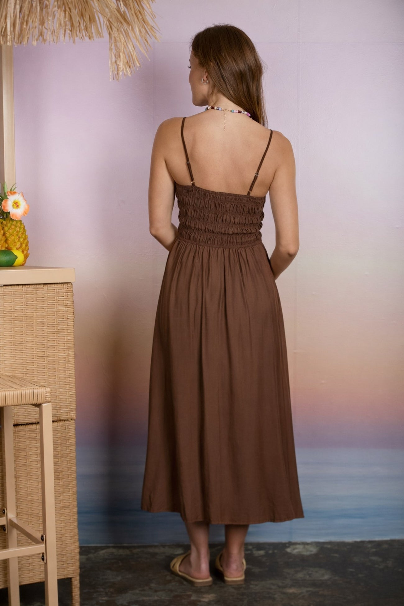woman wearing a brown smocked back maxi dress with thin straps, standing against a pastel background