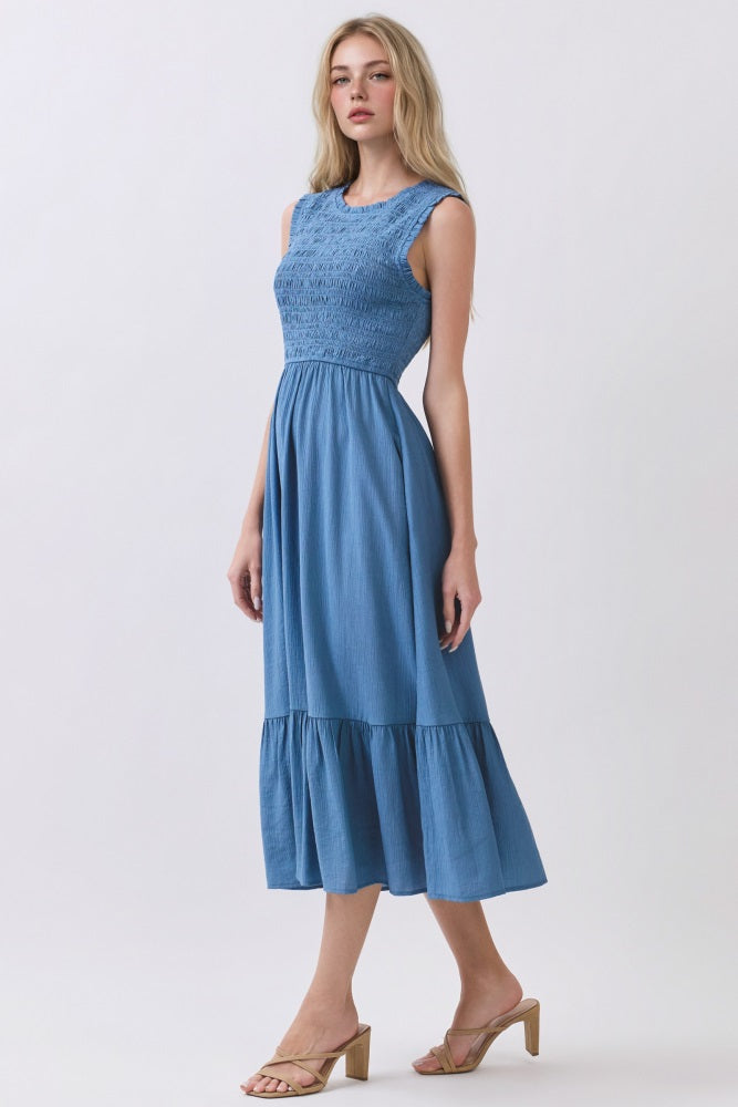 woman wearing a blue sleeveless midi dress with smocked bodice and tiered ruffle hem, standing against a light background