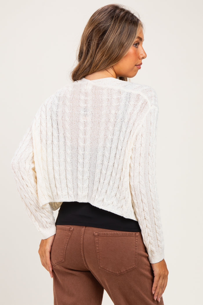 woman wearing a cream cable knit maternity sweater with a cropped back, styled with brown pants