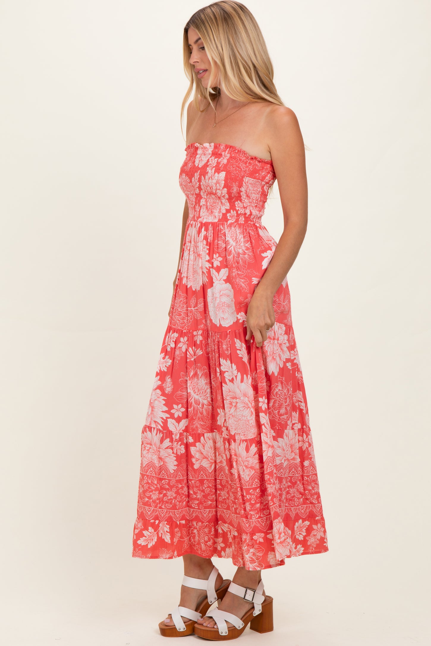 woman wearing a coral strapless floral maxi dress with a fitted bodice and flowing skirt, paired with white block-heel sandals