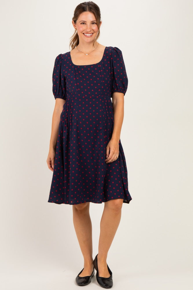 woman wearing a navy blue maternity dress with red polka dots and puffed sleeves, smiling and standing against a neutral background