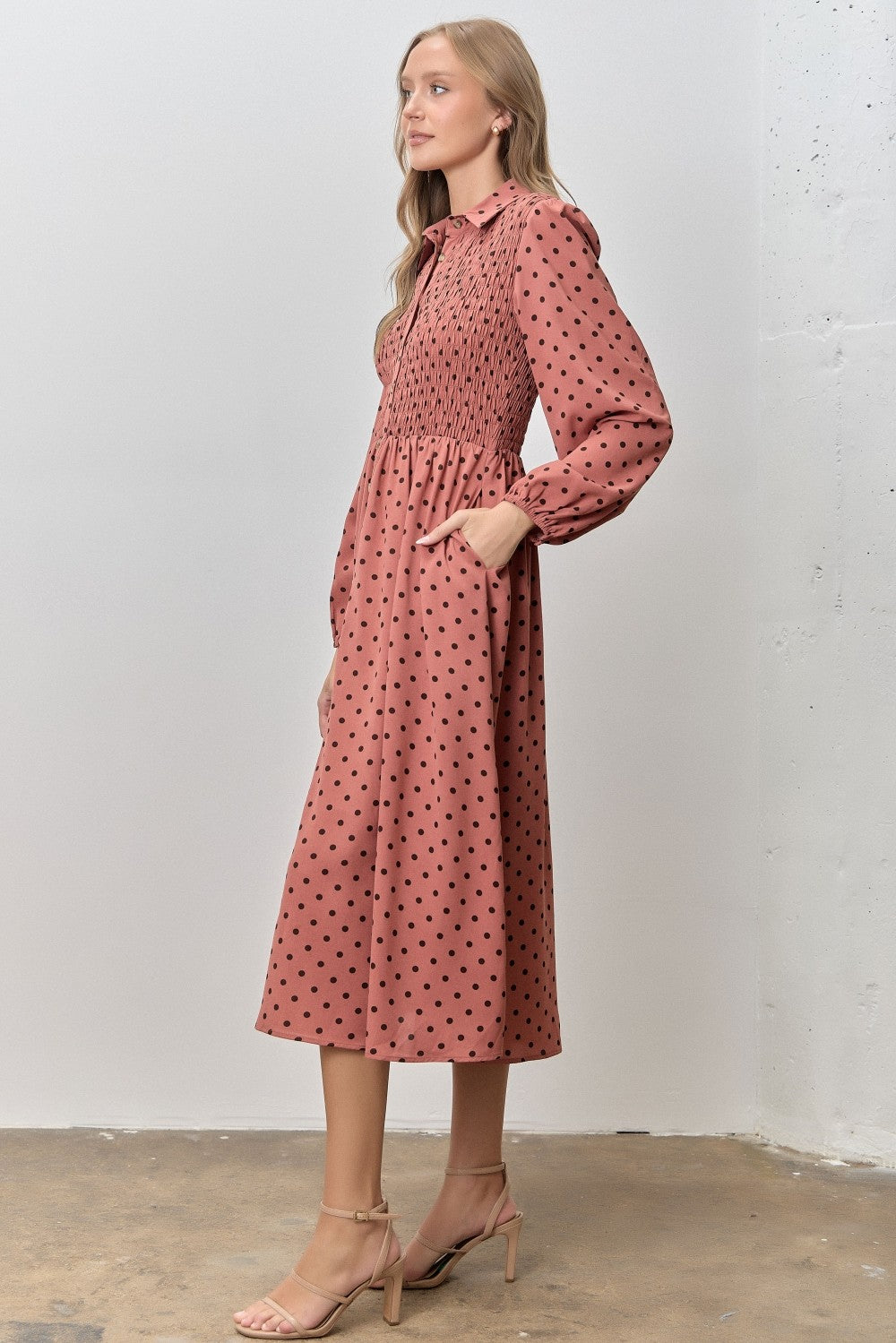 woman wearing a dusty pink long sleeve midi dress with black polka dots and smocked bodice, posing with hands in pockets