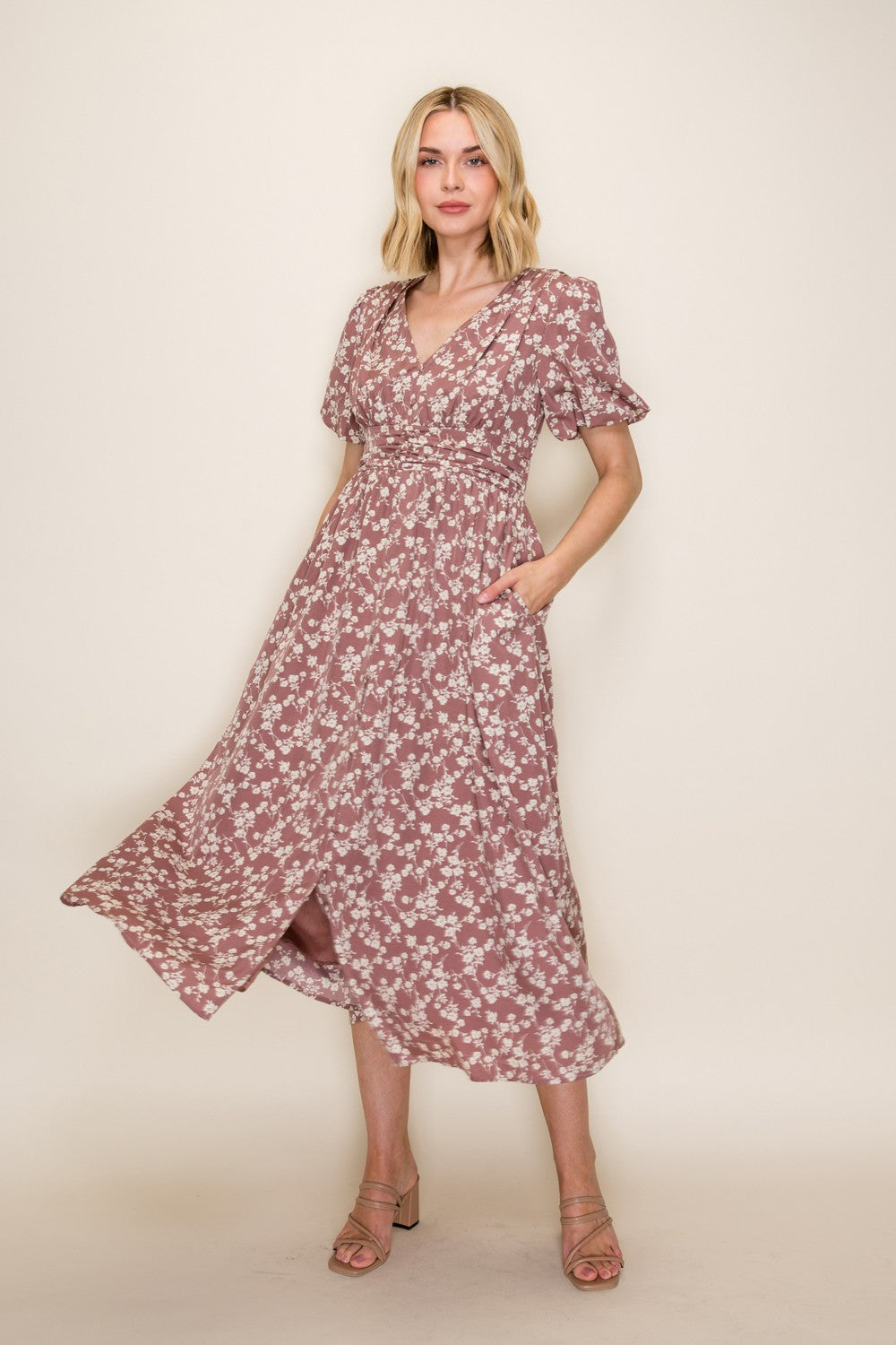 woman wearing a pink floral midi dress with a v-neckline and ruffled sleeves, posing with hands in pockets