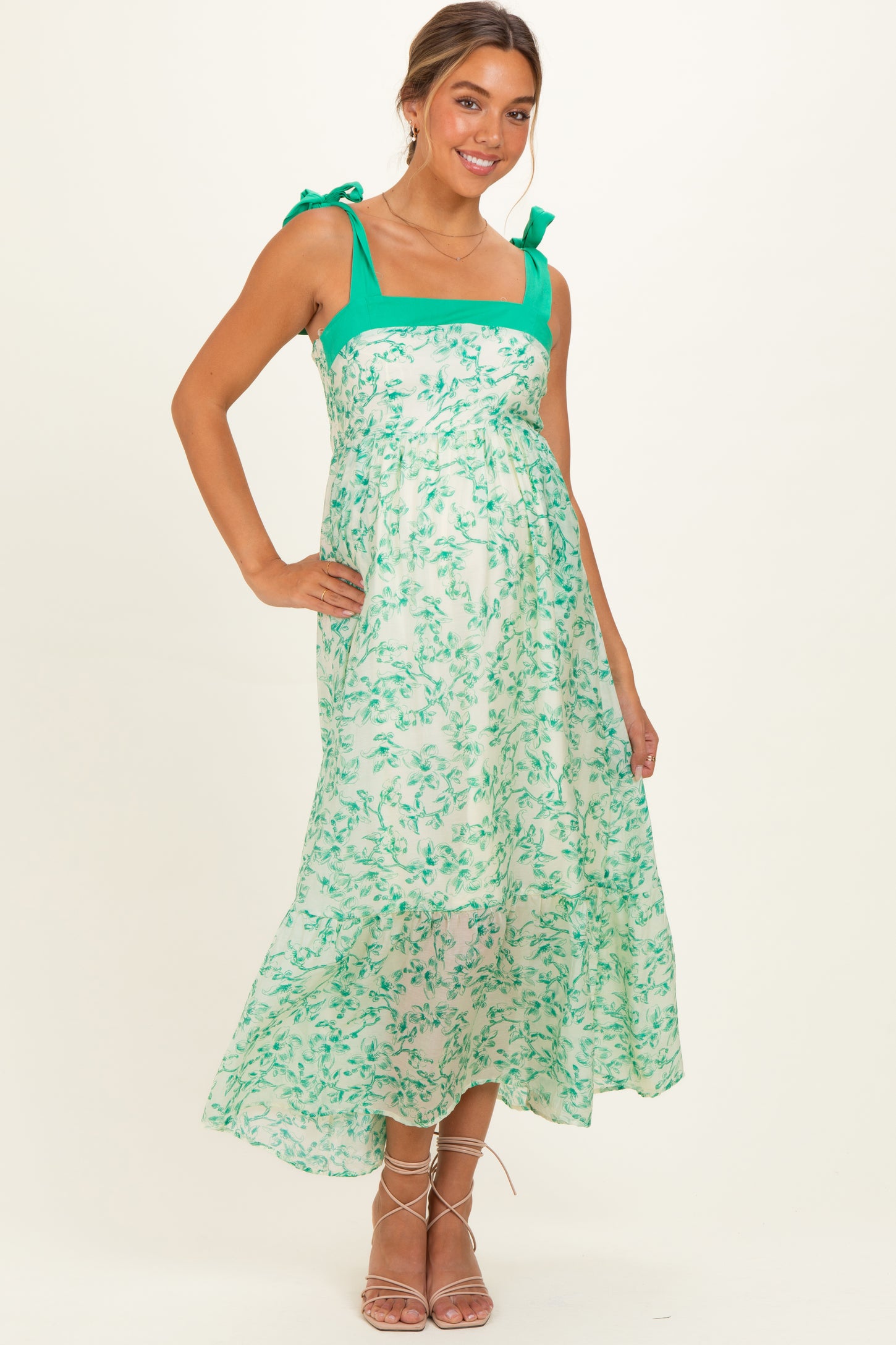 woman wearing a green and white floral maternity dress with adjustable tie straps and a flowing skirt, showcasing her baby bump