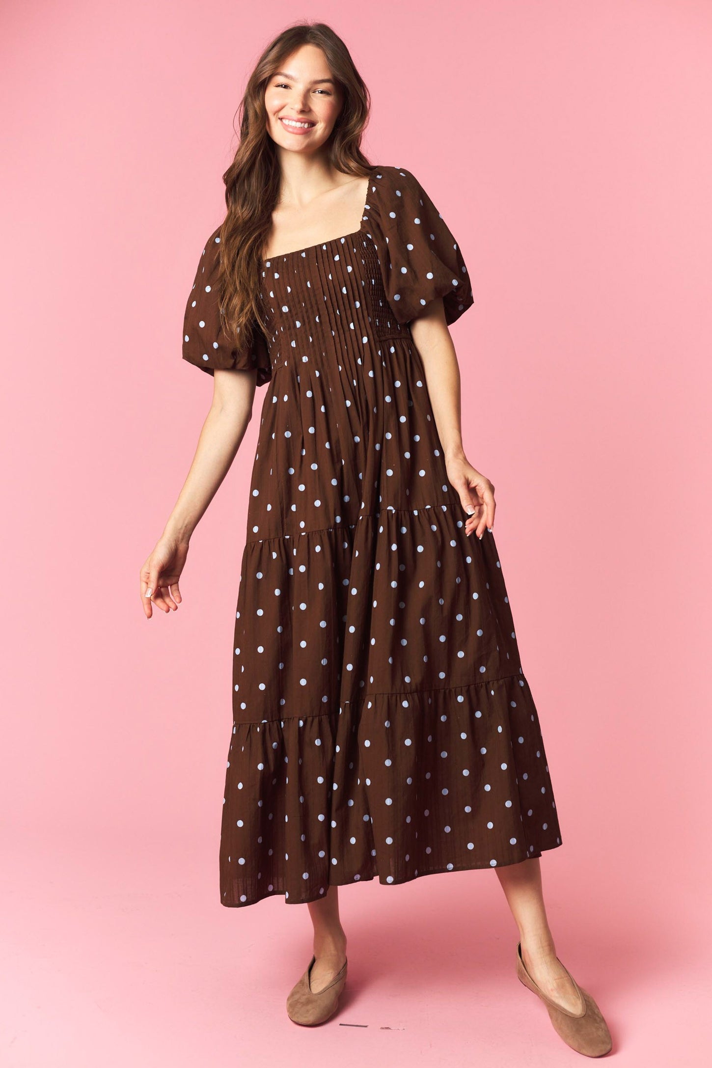 woman wearing a brown polka dot maxi dress with puff sleeves and a tiered skirt standing against a pink background