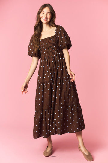 woman wearing a brown polka dot maxi dress with puff sleeves and a tiered skirt standing against a pink background