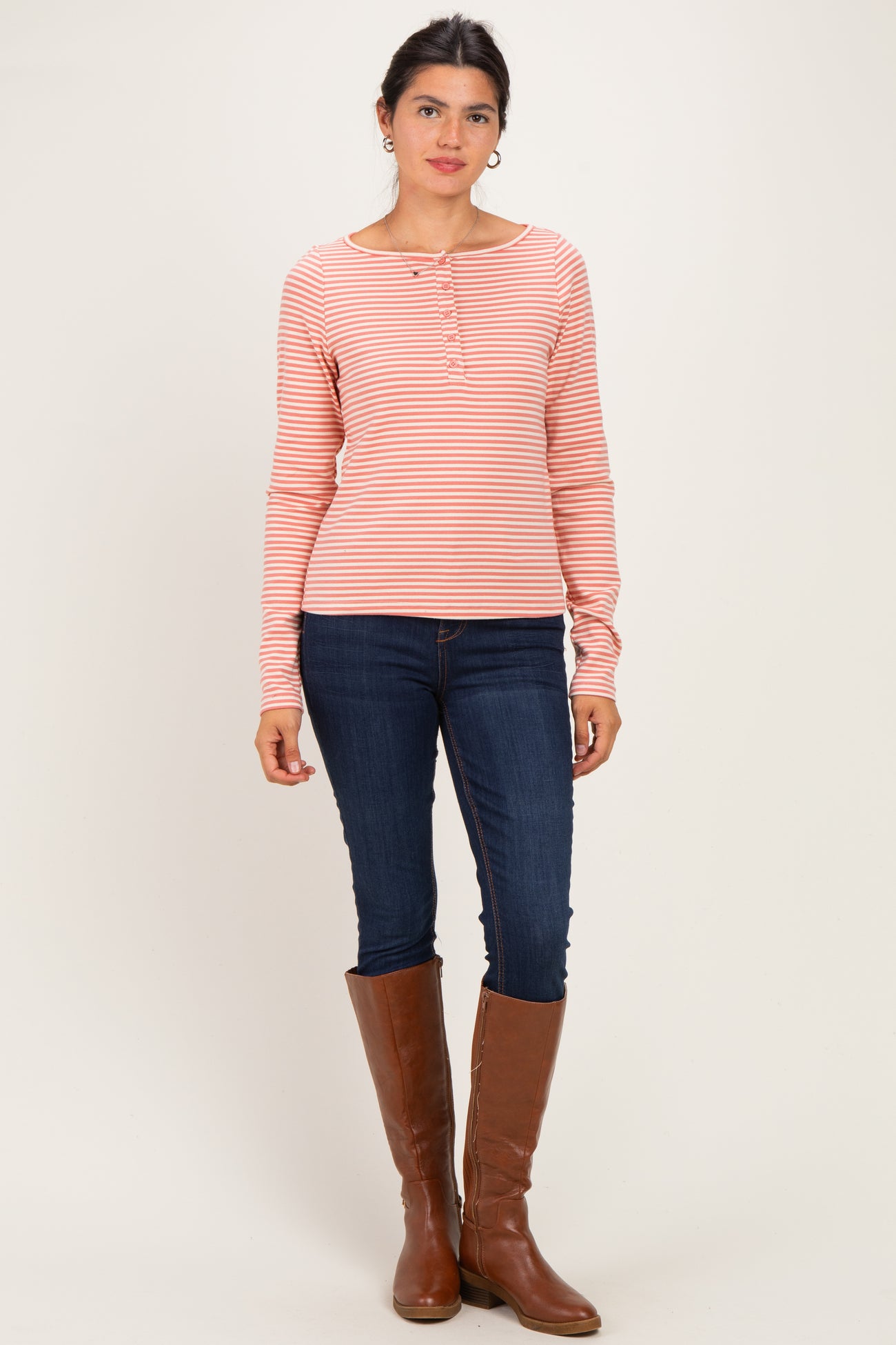 woman wearing a pink and white striped long sleeve henley top with button details and blue skinny jeans, standing in tall brown boots