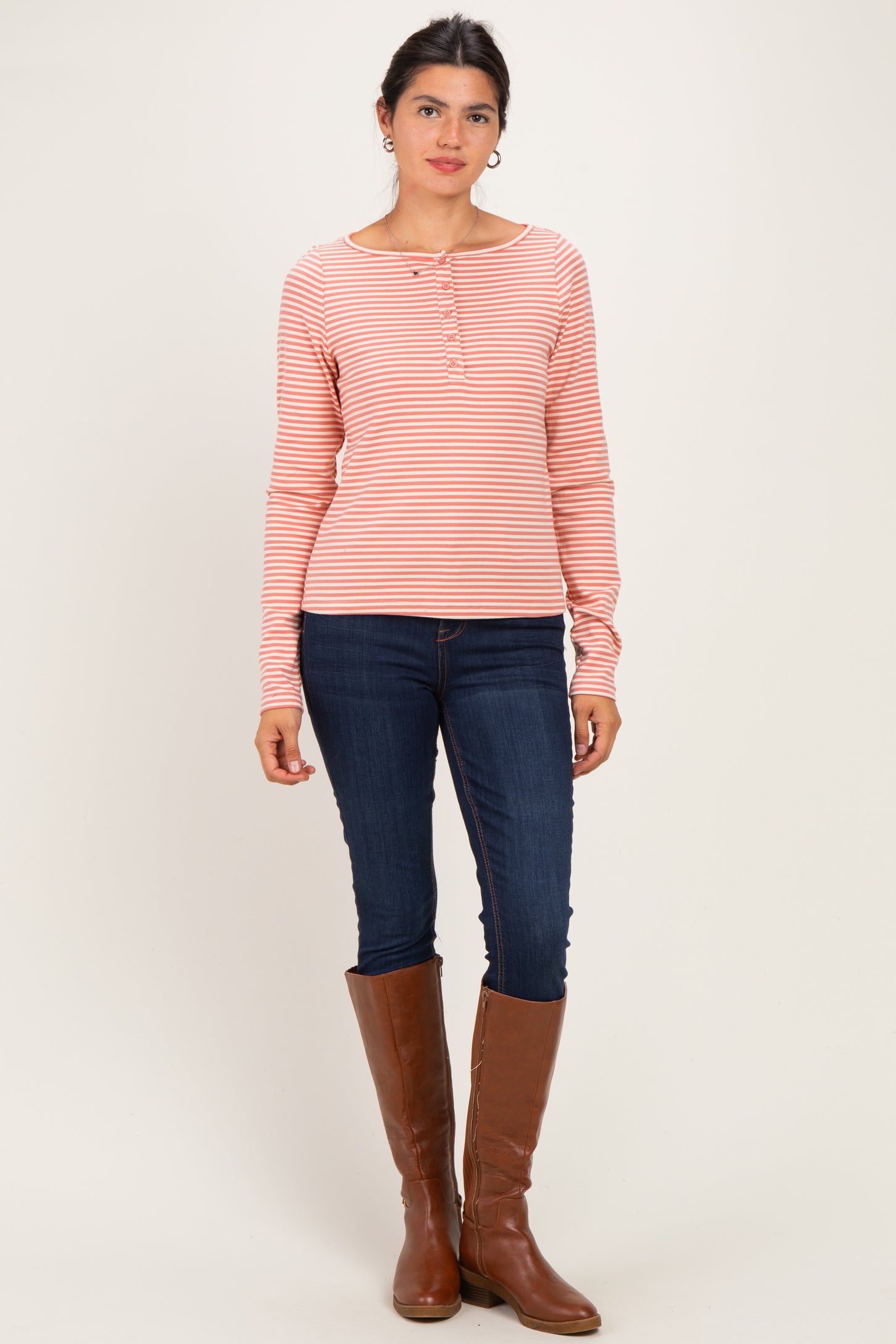 woman wearing a pink and white striped long sleeve henley top with button details and blue skinny jeans, standing in tall brown boots