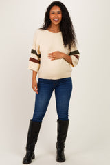 woman wearing a cream maternity sweater with striped sleeves and holding her baby bump, paired with blue jeans and black boots