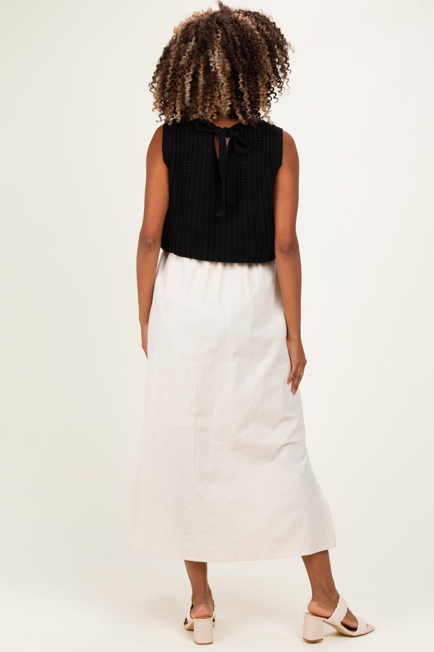 woman wearing a black knitted sleeveless top with a tie detail and a long cream maternity skirt from the back