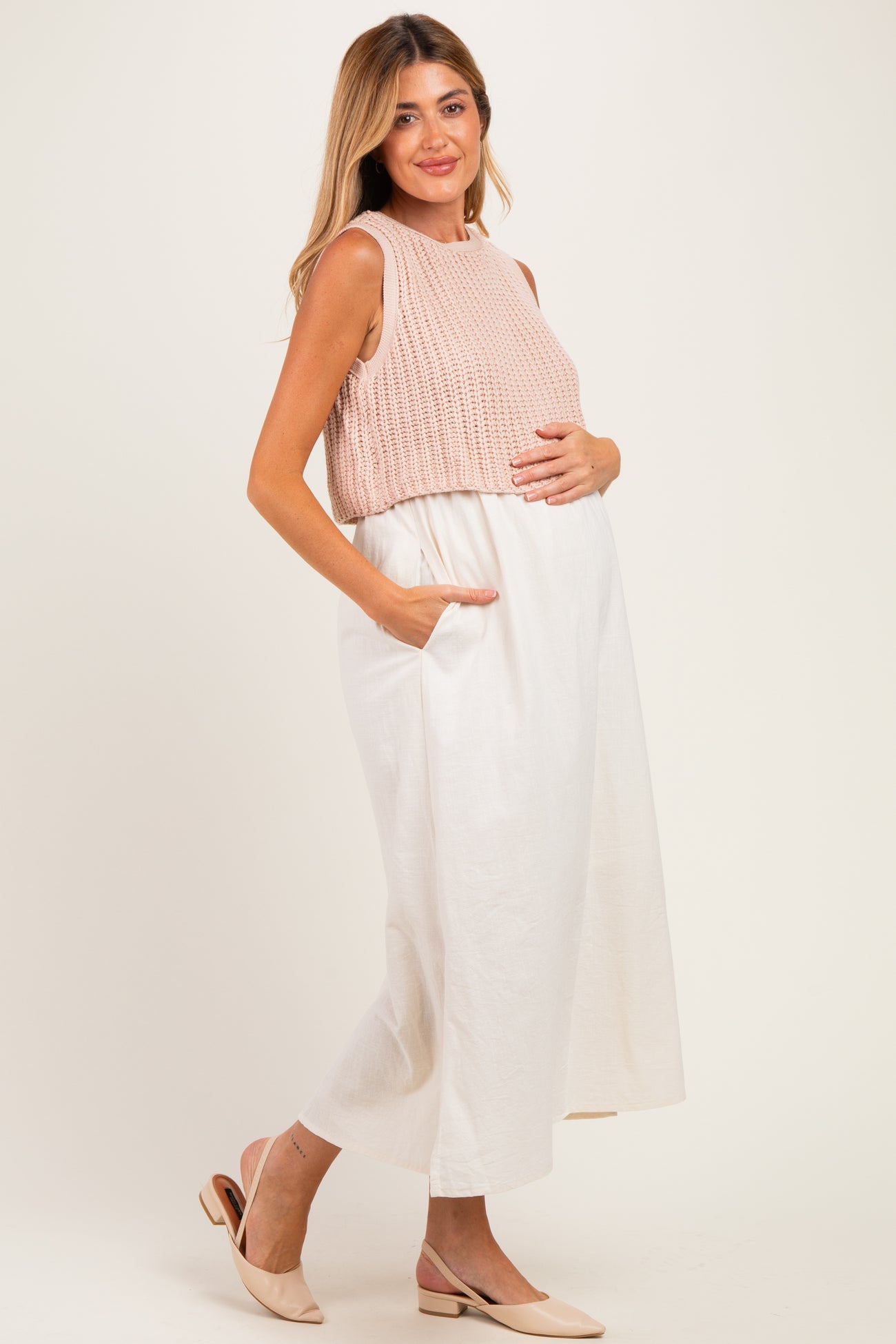 woman wearing a sleeveless peach maternity top and a white maxi skirt, holding her baby bump.