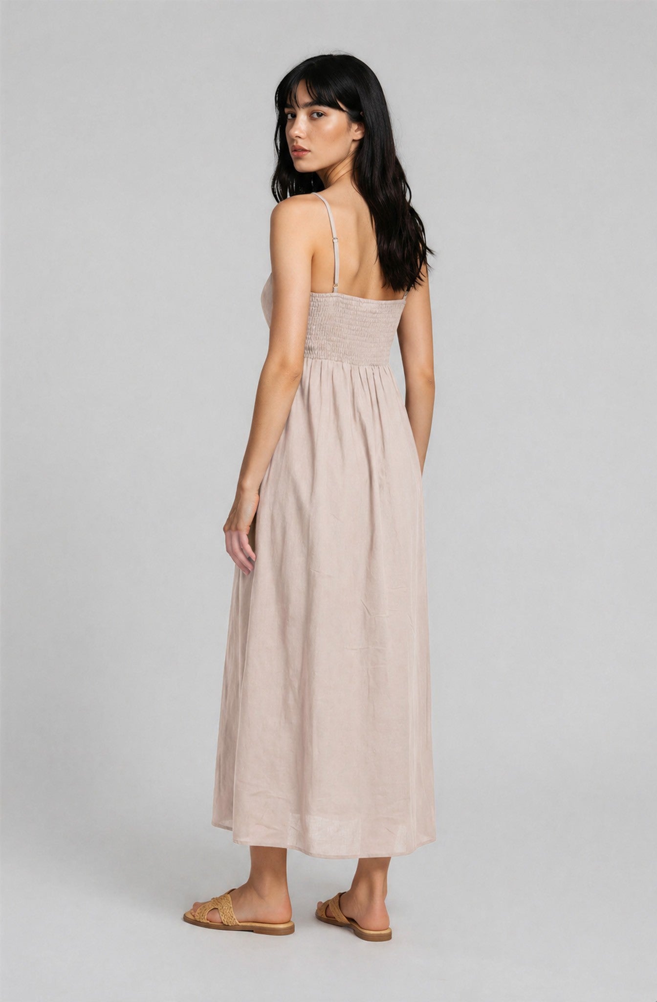 woman wearing a light beige midi dress with a smocked bodice and adjustable straps, standing with her back to the camera