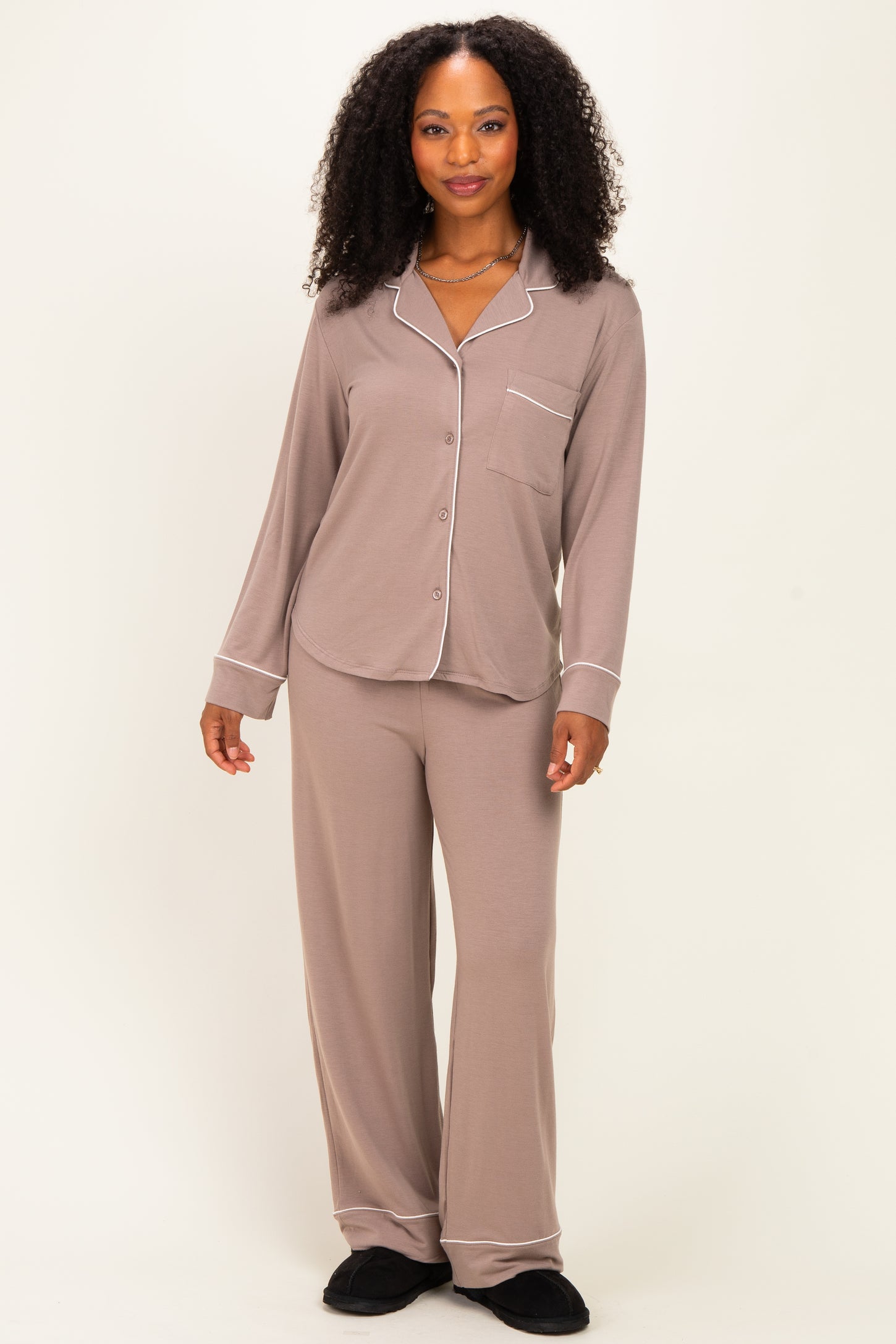 woman wearing a taupe long sleeve pajama set with white piping and matching pants, standing against a neutral background