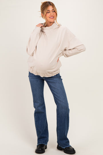 woman wearing a beige maternity sweatshirt with a high neck and a visible baby bump, paired with blue flared jeans