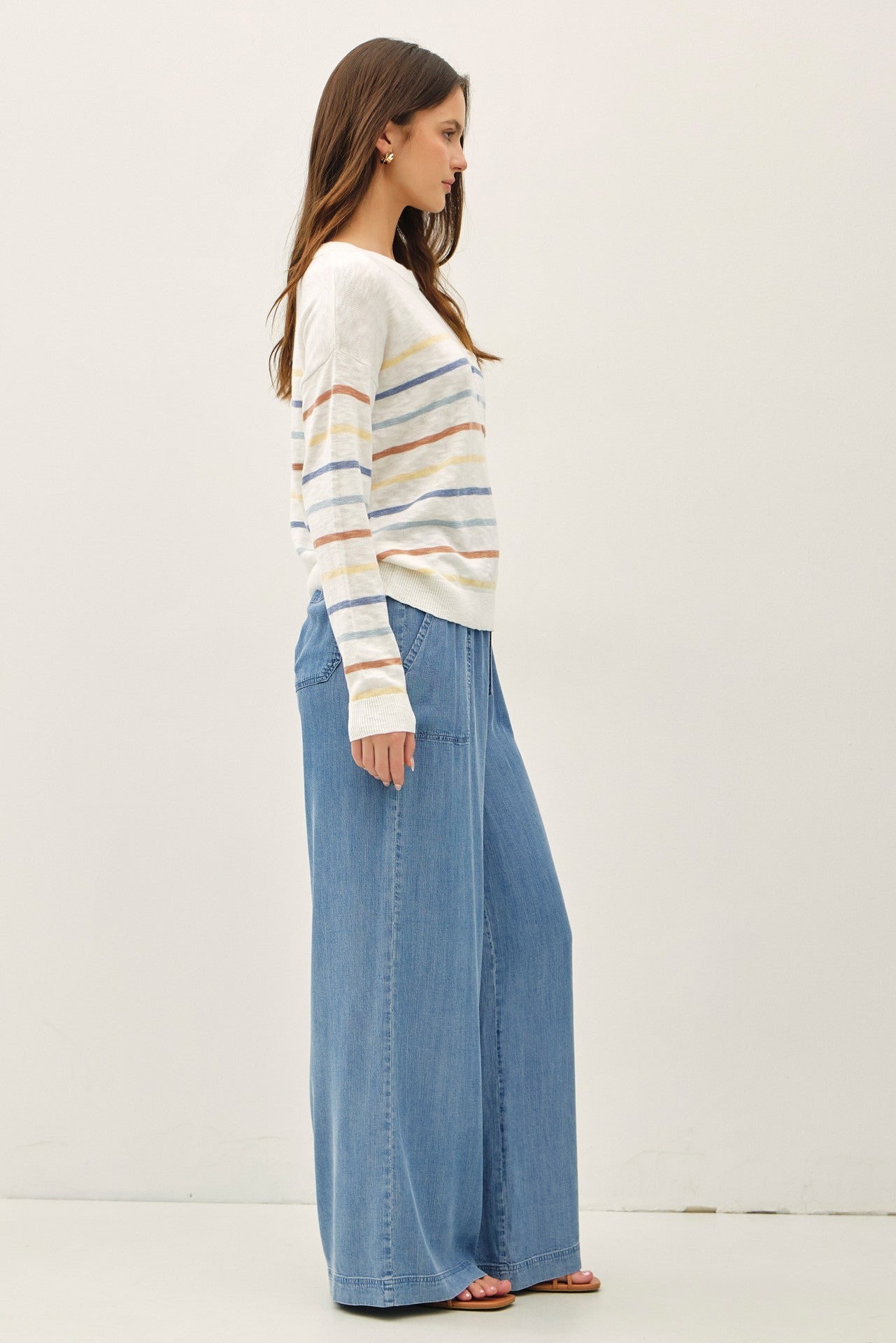 woman wearing a cream striped sweater and wide-leg blue denim pants, standing in profile against a white background