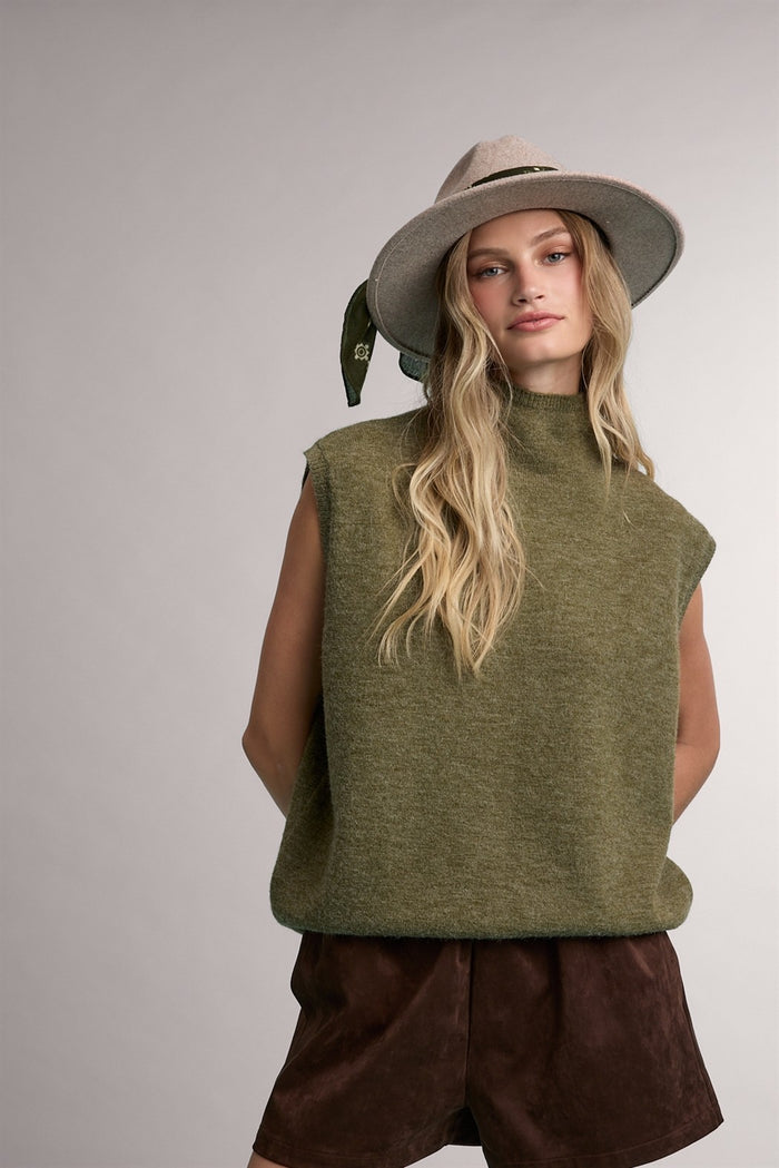 woman wearing an olive green sleeveless sweater with a high neck and brown suede shorts, styled with a hat and scarf