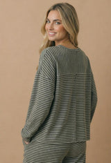 woman wearing a gray and white striped long sleeve top, smiling over her shoulder against a neutral background