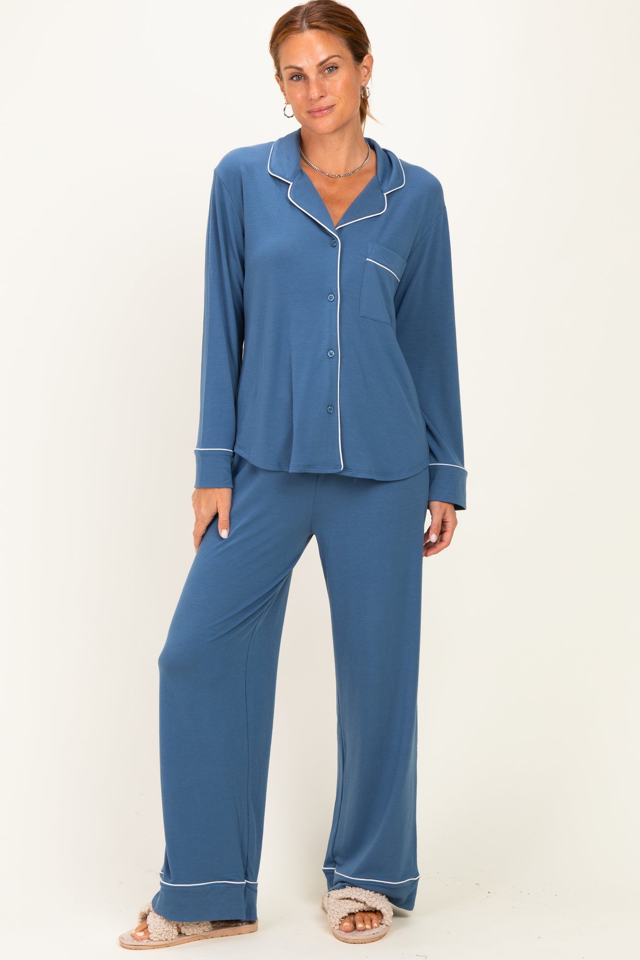 woman wearing a blue pajama set with long sleeves, pants, and white piping details, standing against a plain background