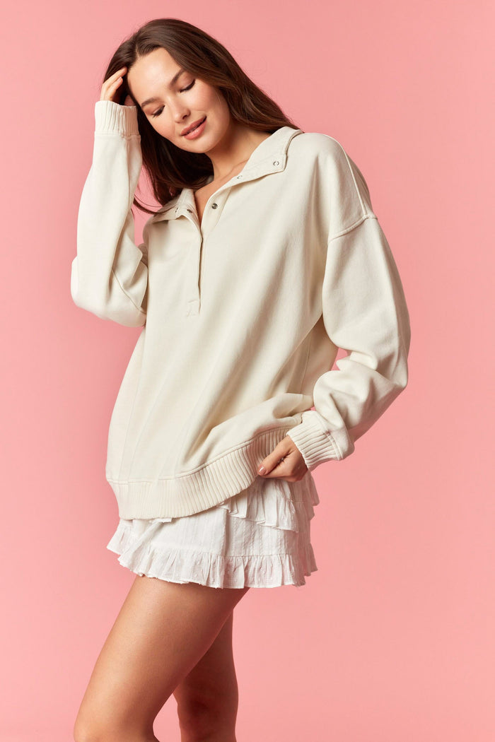 woman wearing a cream oversized quarter-zip sweatshirt with a layered ruffle skirt against a pink background